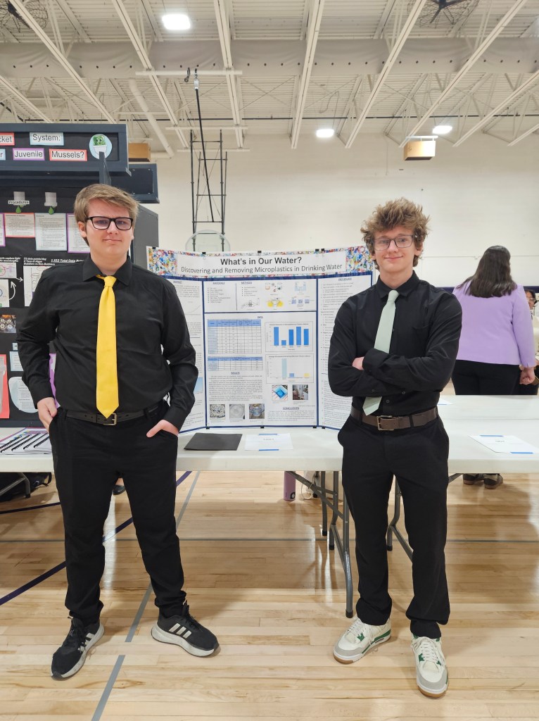 Two students dressed in formal attire stand in front of a science fair project poster titled 'What's in Our Water? Discovering and Removing Microplastics in Drinking Water.' The poster includes graphs and images related to the project, displayed in a gymnasium setting.