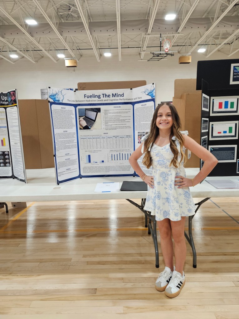 A young girl in a floral dress stands beside a science project display titled 'Fueling The Mind,' which examines the link between hydration levels and cognitive performance, in a school gymnasium.