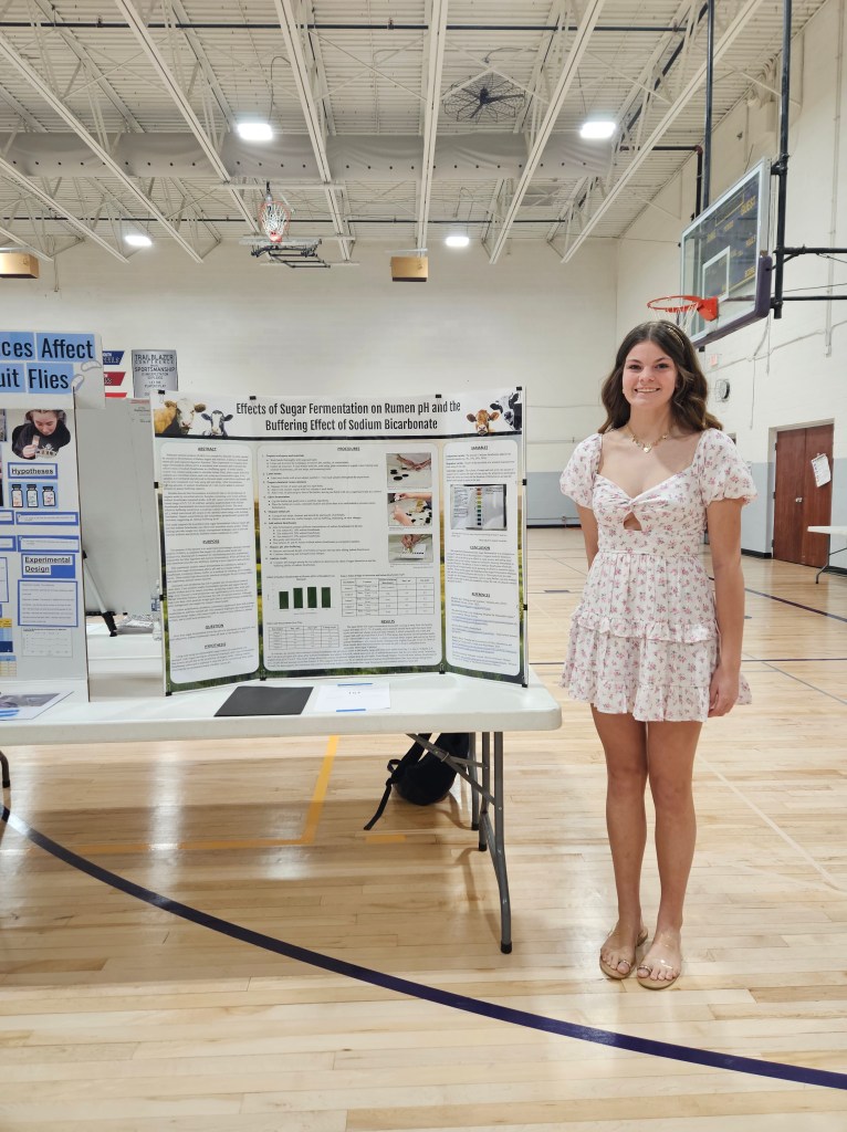 A young woman in a floral dress stands next to a science poster presentation titled 'Effects of Sugar Fermentation on Rumen pH and the Buffering Effect of Sodium Bicarbonate' in a gymnasium.