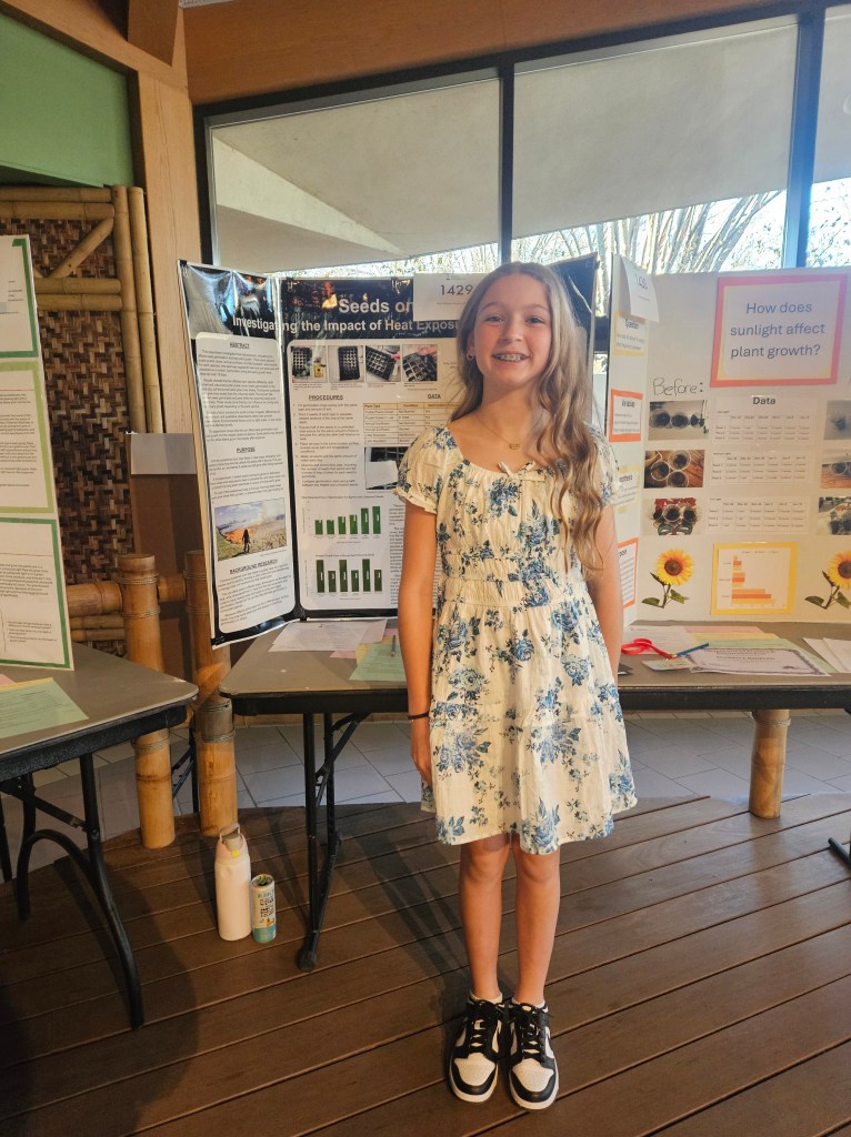 A young girl wearing a floral dress stands in front of science project displays, smiling confidently. The projects focus on investigating the impact of heat exposure on seeds and how sunlight affects plant growth.