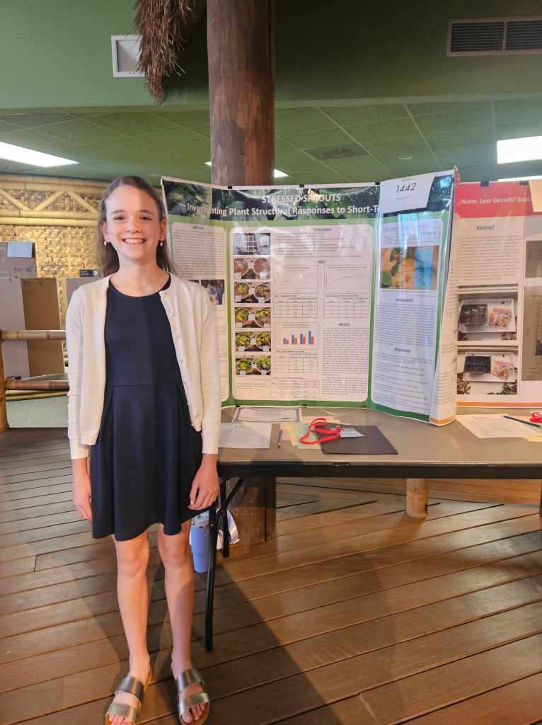A young girl stands smiling in front of a science fair project display titled 'Stressed Sprouts: Investigating Plant Structural Responses to Short-Term Stress'. The poster includes graphs and images related to the project, and a table is in front holding additional materials.