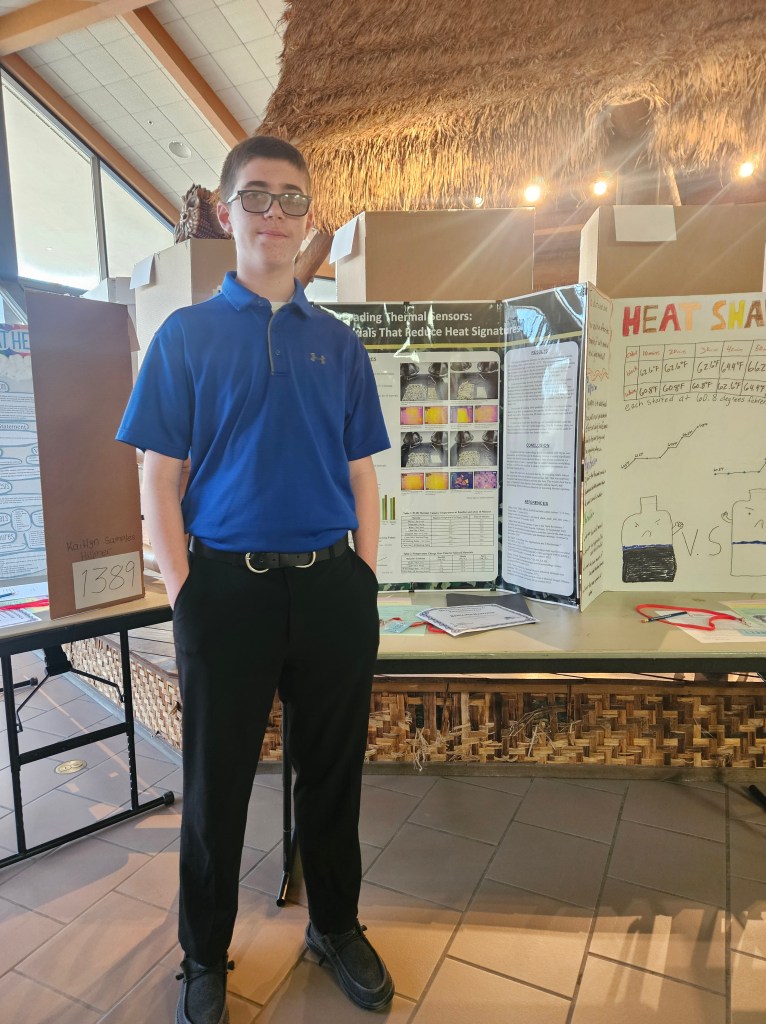 A student standing in front of a science project display at a fair, wearing a blue shirt and black pants, with various project posters visible in the background.