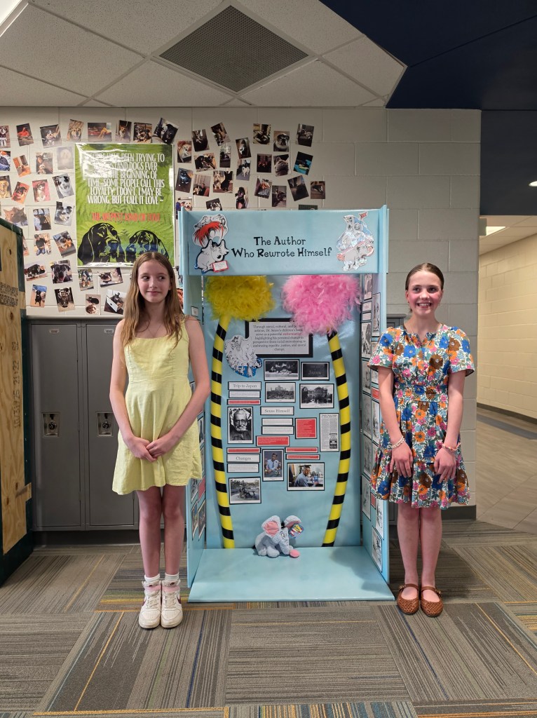 Two girls standing in front of a display titled 'The Author Who Rewrote Himself' decorated with colorful feathers and photographs, in a school setting.