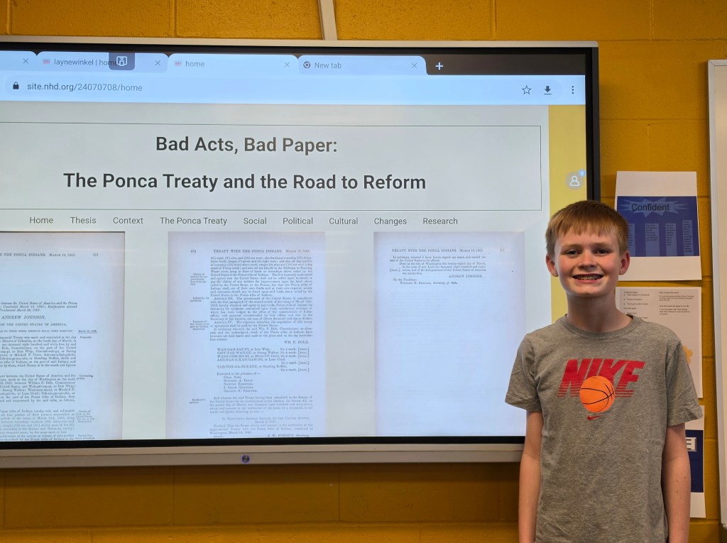 A young boy stands next to a presentation on a screen, which reads 'Bad Acts, Bad Paper: The Ponca Treaty and the Road to Reform.' The screen displays text and images related to the Ponca Treaty. The boy wears a gray Nike t-shirt and has a friendly smile.