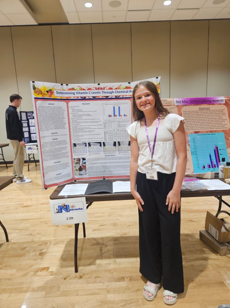 A young girl stands next to a science project display board titled 'Determining Vitamin C Levels Through Chemical Analysis' at a science fair. She is wearing a white top and black pants, smiling at the camera.