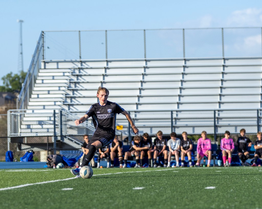 A young player in a black soccer uniform kicking a soccer ball on a field, with seated teammates in the background.