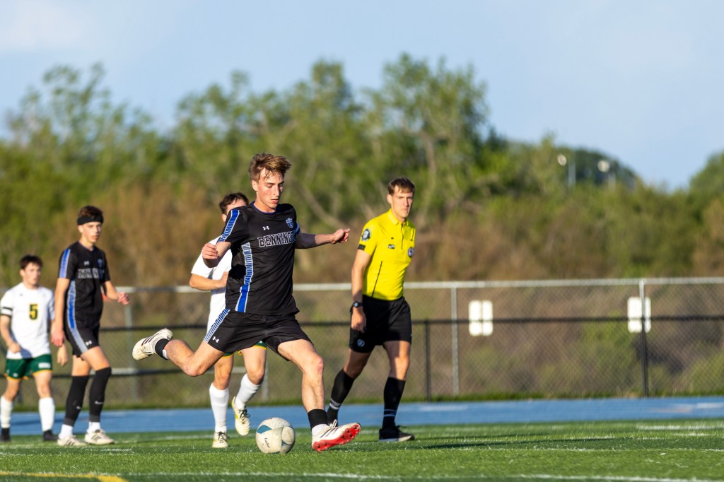 A soccer player in a black jersey with blue accents kicks a ball on the field, with other players and a referee in the background.