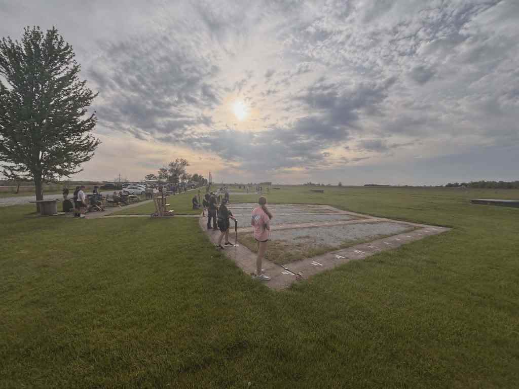 A cloudy sky with a sunset, people gathered on a grassy field, some sitting and others standing near a measurement area.