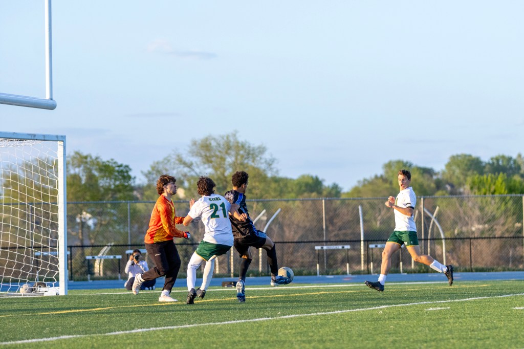 A soccer match in progress with players competing for the ball near the goal. A goalkeeper in an orange jersey defends, while other players in green and white uniforms are actively involved in the play on a grassy field.