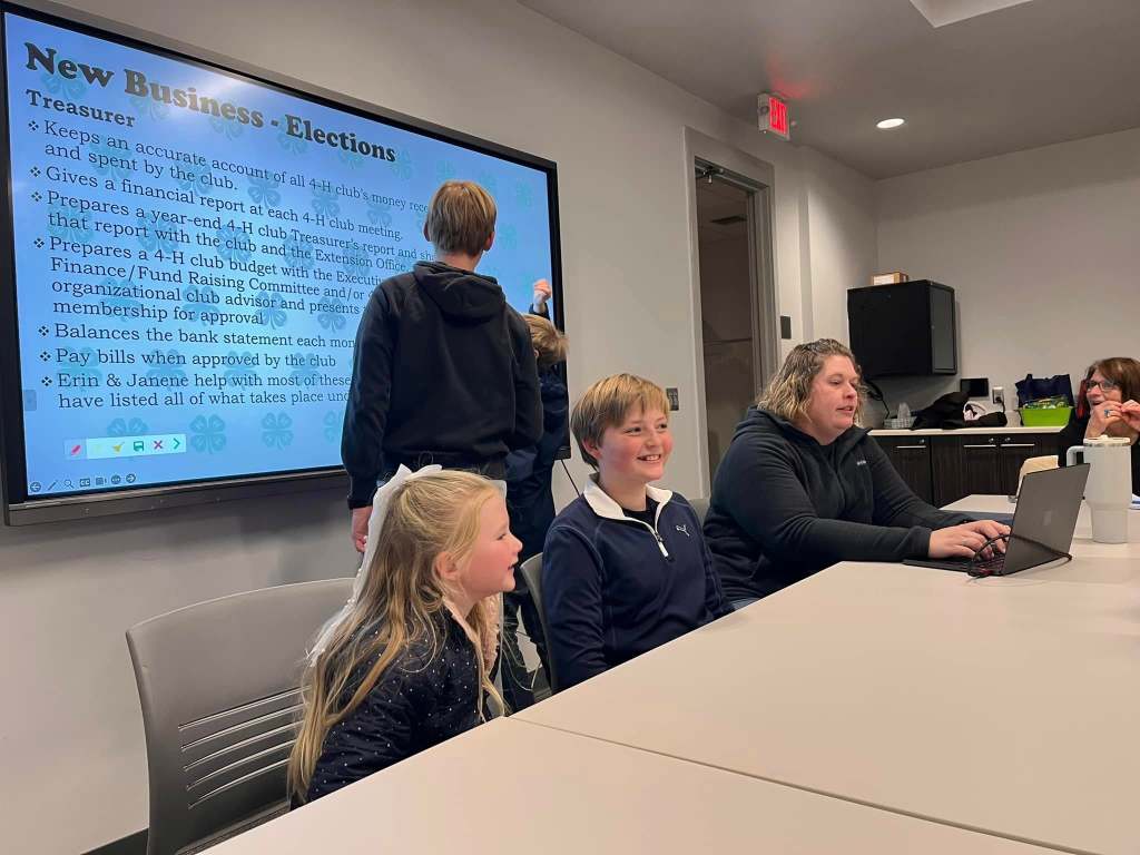 A group of people attending a 4-H meeting, with children and adults seated at a table. A screen displays information about club elections and treasurer responsibilities. One child is sitting in front of the screen while another stands in the background.