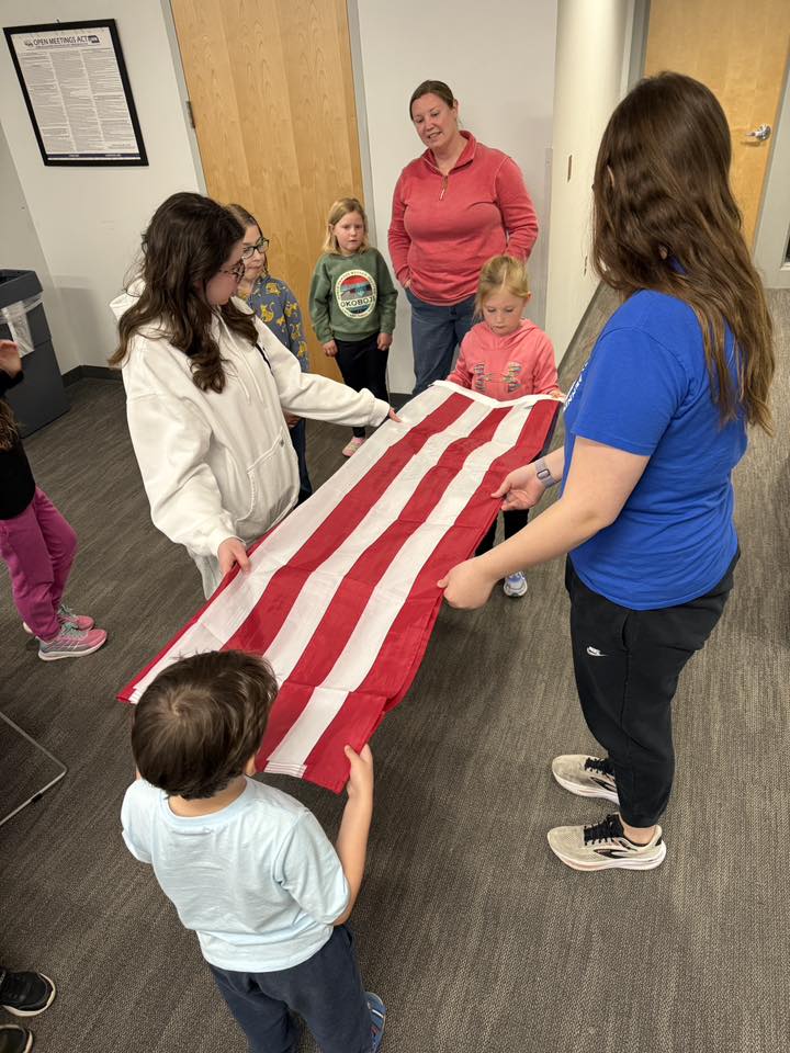 A group of children and adults are gathered indoors, holding an American flag. The adults are guiding the children in a collaborative activity involving the flag.