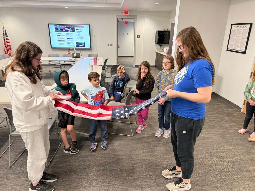 A group of children and teens are gathered in a room, with some sitting and others standing. One girl in a blue shirt is holding an American flag while another girl in a white jacket is assisting her. The background includes a screen displaying information and flags.