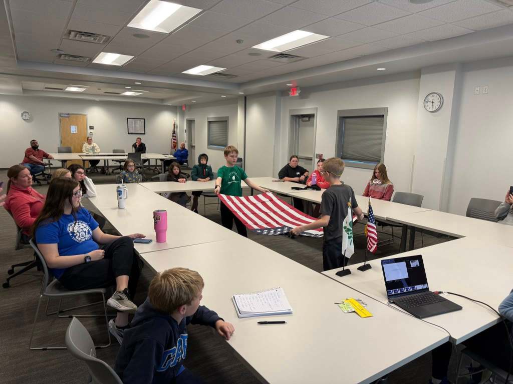 A group of children and adults gathered in a meeting room, with two boys presenting a folded American flag. The audience, seated at tables, watches attentively.