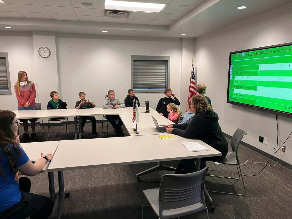 A group of children and adults seated around a table in a meeting room, with a large screen displaying a financial report. A girl stands in the background while others are focused on the presentation. The American flag is visible in the corner.