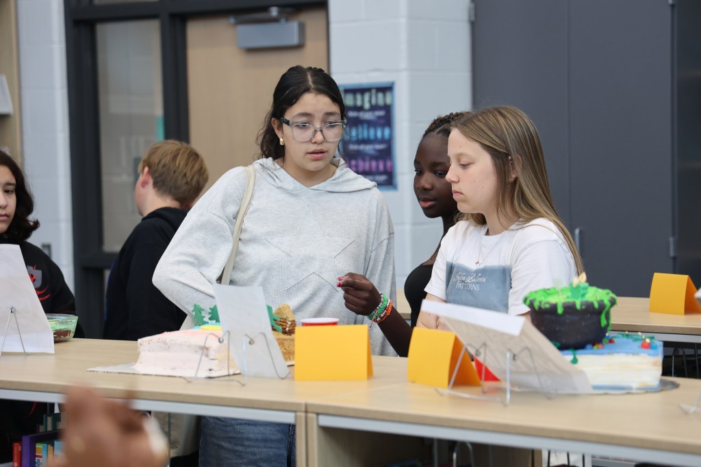 Students observing and discussing decorated cakes at a school event, with various cake designs displayed on tables.