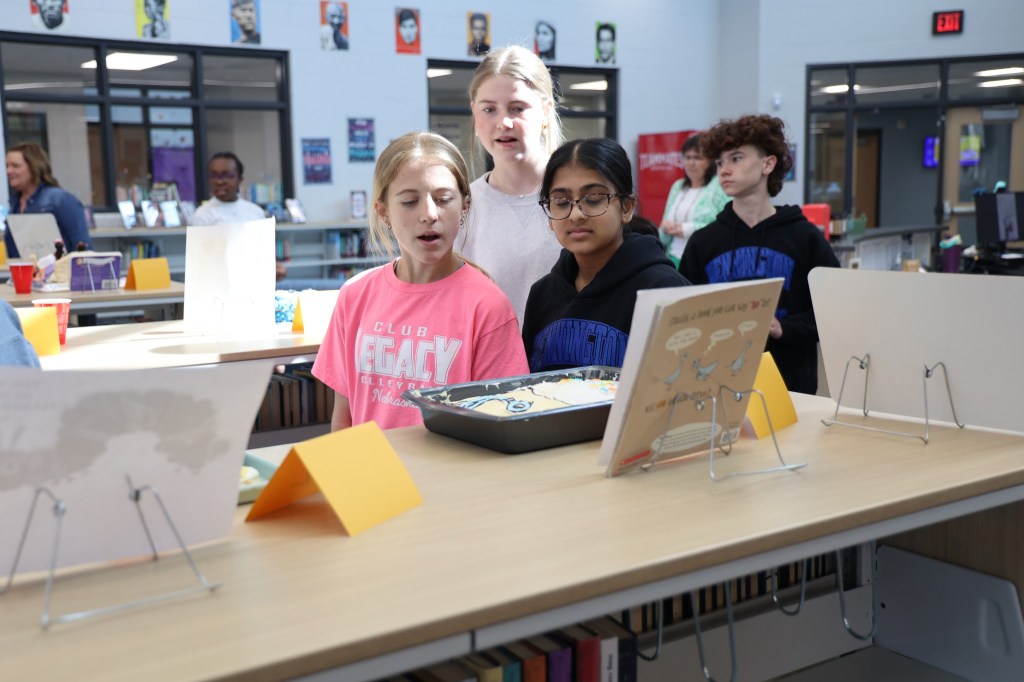 Students participating in an event at a library, observing a dessert display on a table while others look on.