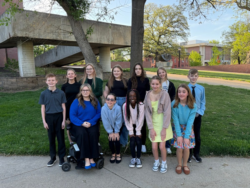 A group of twelve children and teens posing together outdoors, with some sitting and others standing, in a grassy area near a modern building and trees.