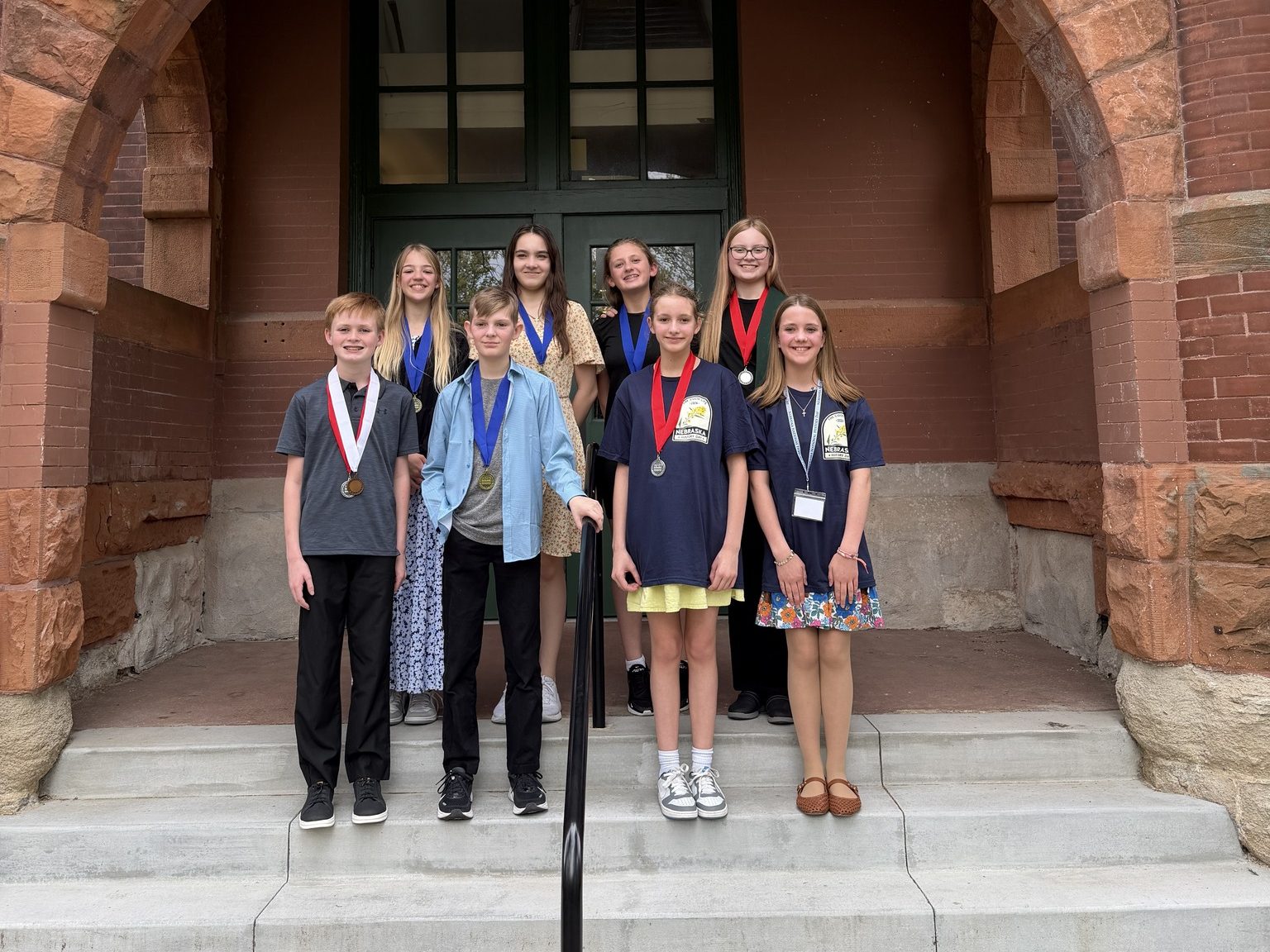 A group of eight students standing on the steps of a building, all wearing medals. They are smiling and dressed in casual attire, with some wearing t-shirts and dresses. The background features a historical brick building with large green doors.