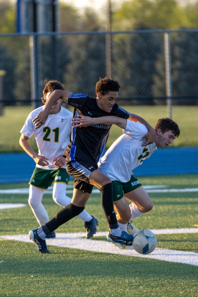 A soccer player in a black jersey competes for the ball against an opponent in a white jersey, while two other players are in the background on a grassy field.