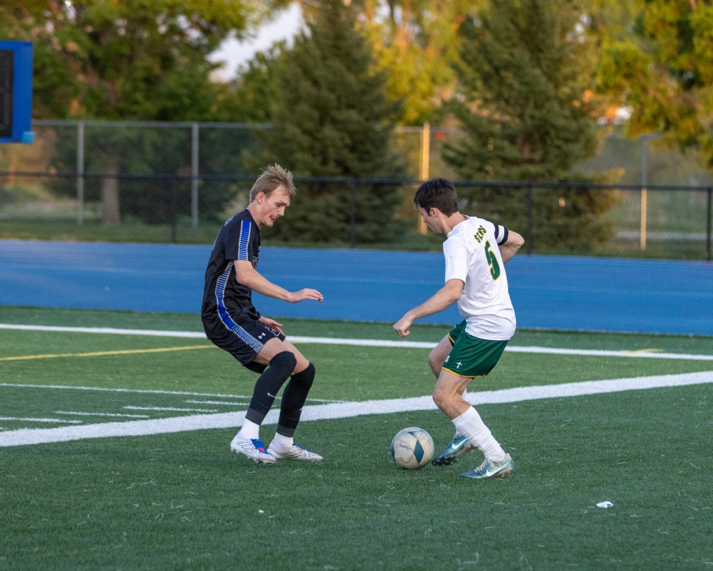 Two soccer players engaging in a game on a field, one in a black jersey and the other in a white jersey with green shorts, dribbling a soccer ball.