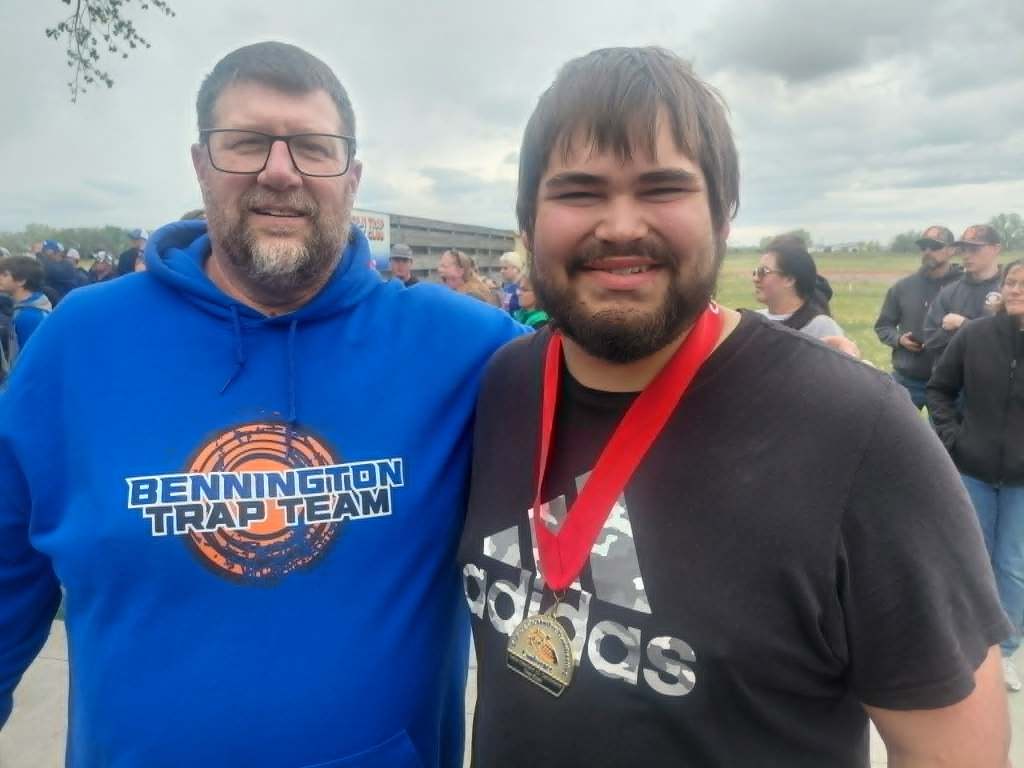 Two men standing together outdoors, smiling. One man wears a blue hoodie with 'Bennington Trap Team' printed on it, and the other wears a black shirt with an adidas logo and a medal hanging from his neck.