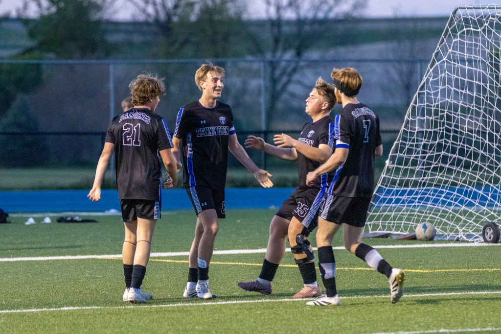 A group of four young soccer players celebrating a goal on the field at dusk, wearing black jerseys with blue accents. One player shows excitement, while another raises his arms in joy.