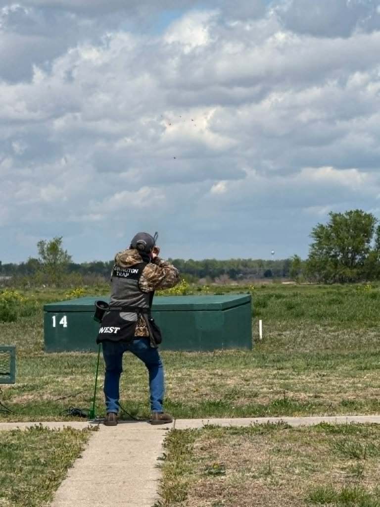 A shooter stands on a shooting range, aiming at flying targets in the sky, with green grass and a cloudy sky in the background.