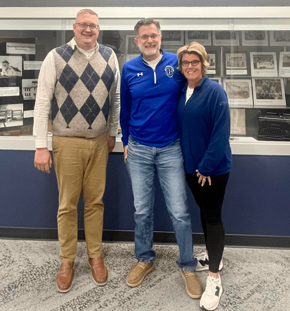 Three individuals standing together in front of a display case, smiling. One person is wearing a patterned sweater vest, another is in a blue sports jacket, and the third is in a blue sweater. The background features black and white photographs.