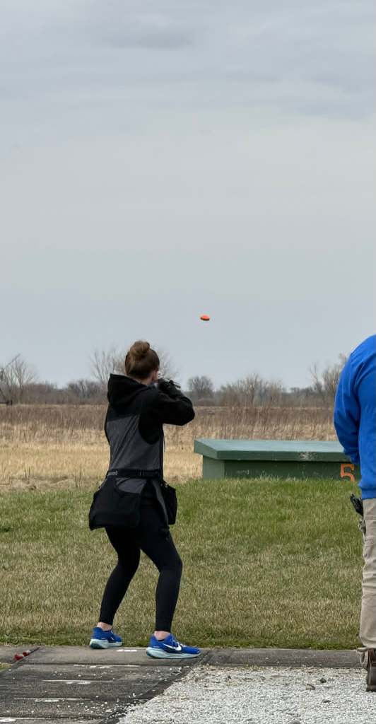 A person in a shooting stance aiming at a clay target in the sky at an outdoor shooting range.