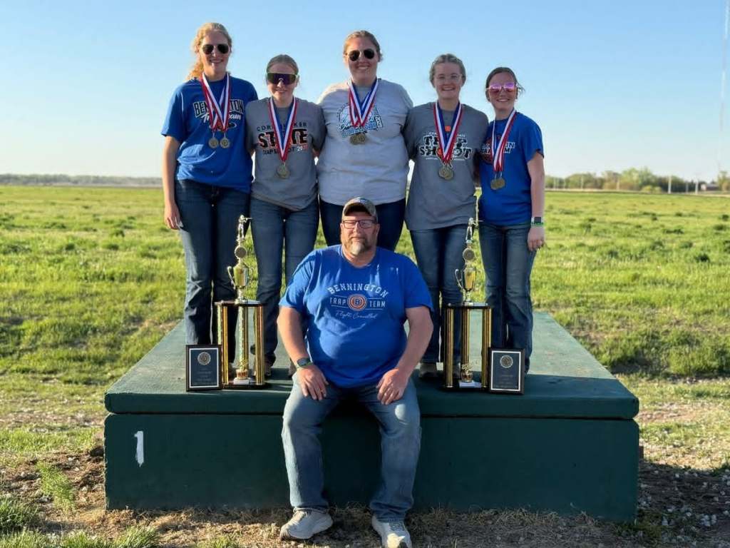 A group of six individuals, including five young women wearing medals and t-shirts, posing on a platform with trophies in a grassy field under a clear sky.