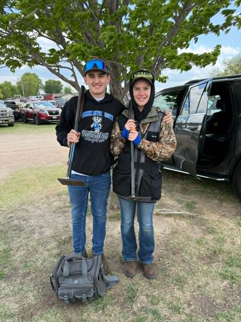 Two young individuals standing outdoors in front of trees and parked vehicles, holding tools, with one wearing a black hoodie and the other in a camouflage jacket.