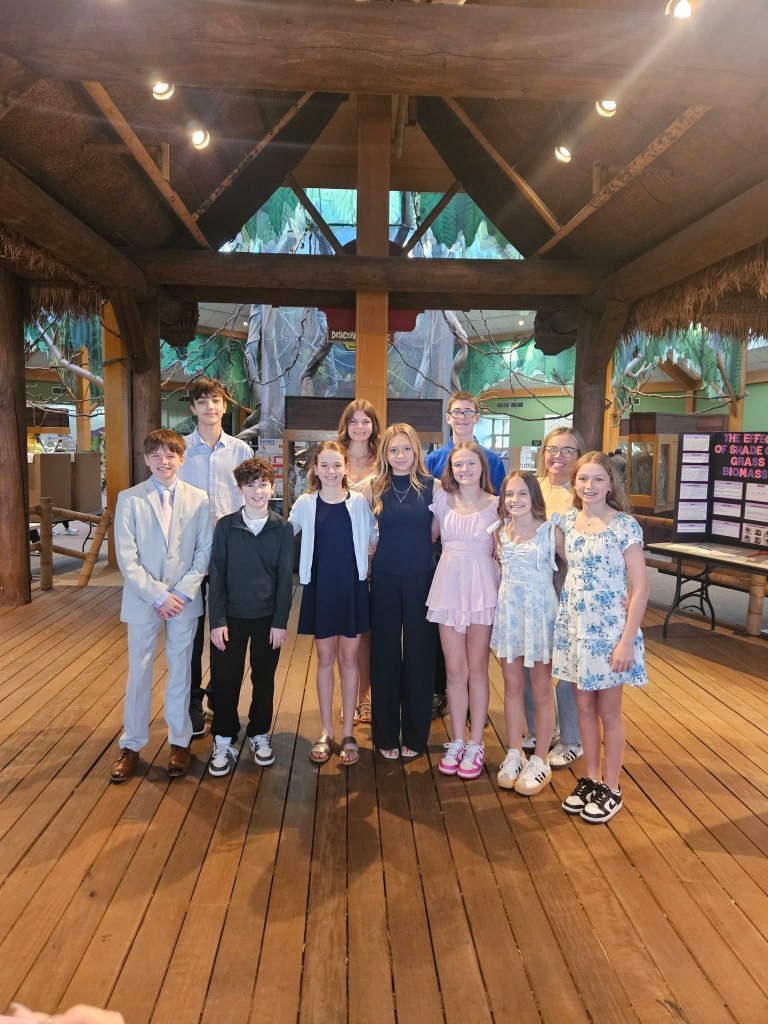 A group of twelve children and teens standing together in a decorated indoor setting, smiling at the camera. The background features wooden beams and greenery, suggesting a tropical theme.