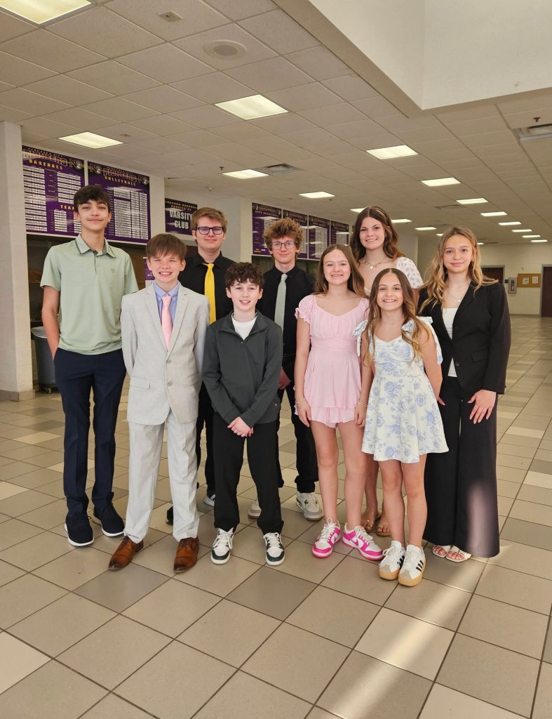 Group of nine young people in formal attire posing together in a school hallway.
