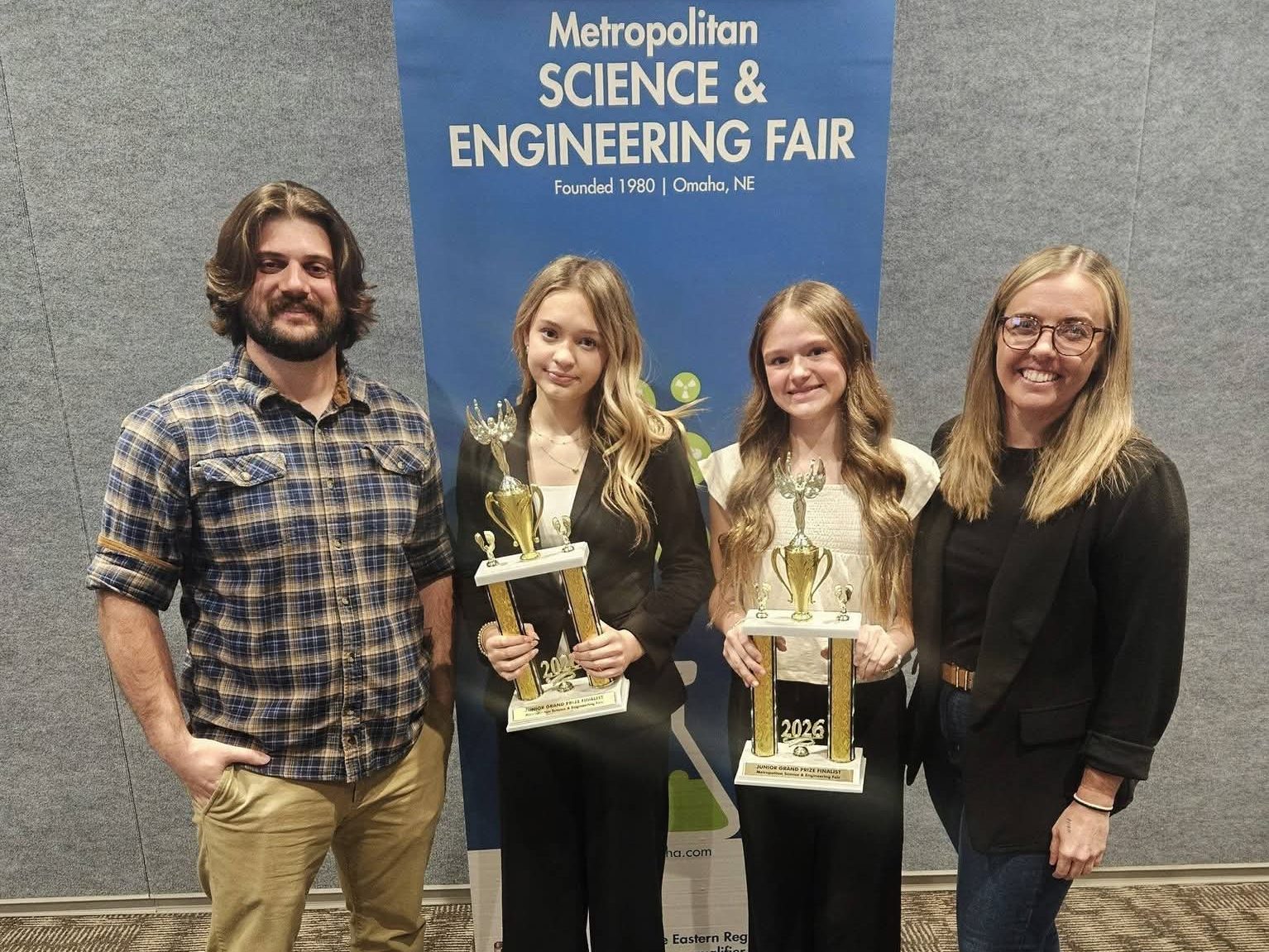 Four individuals stand in front of a banner for the Metropolitan Science & Engineering Fair. Two young girls hold trophies for their achievements, while a man and a woman stand beside them, smiling.