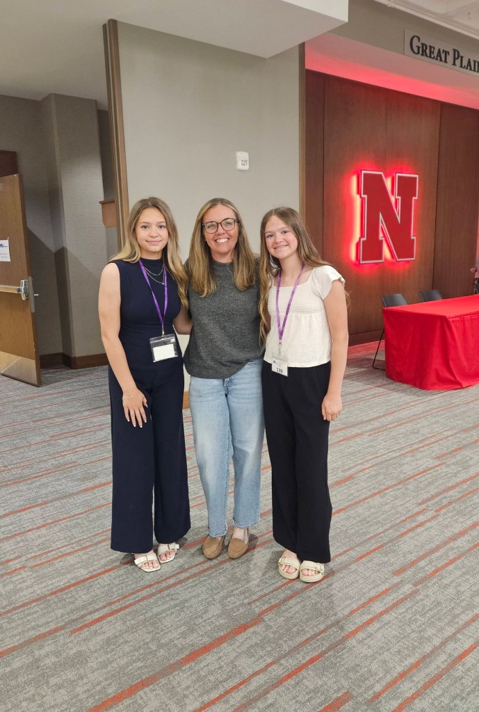Three women posing together indoors, two wearing dark and light-colored outfits and the third wearing a gray top and jeans, with a large Nebraska 'N' sign in the background.
