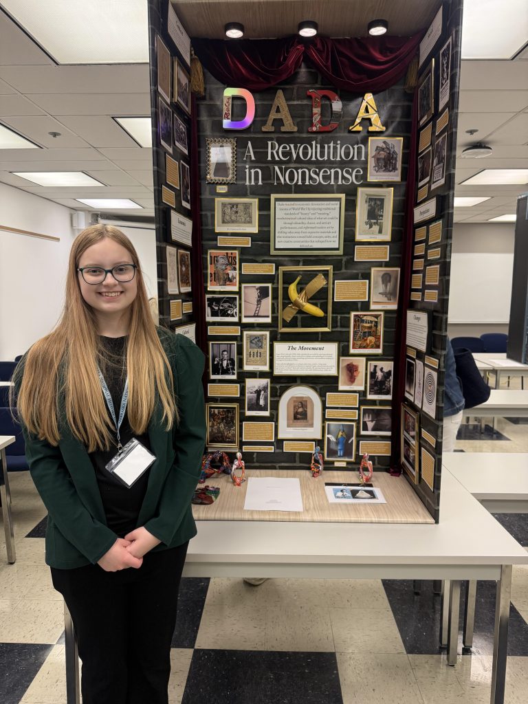 A student presenting a Dada art project titled 'A Revolution in Nonsense,' featuring a display board with various images and text about the Dada movement.