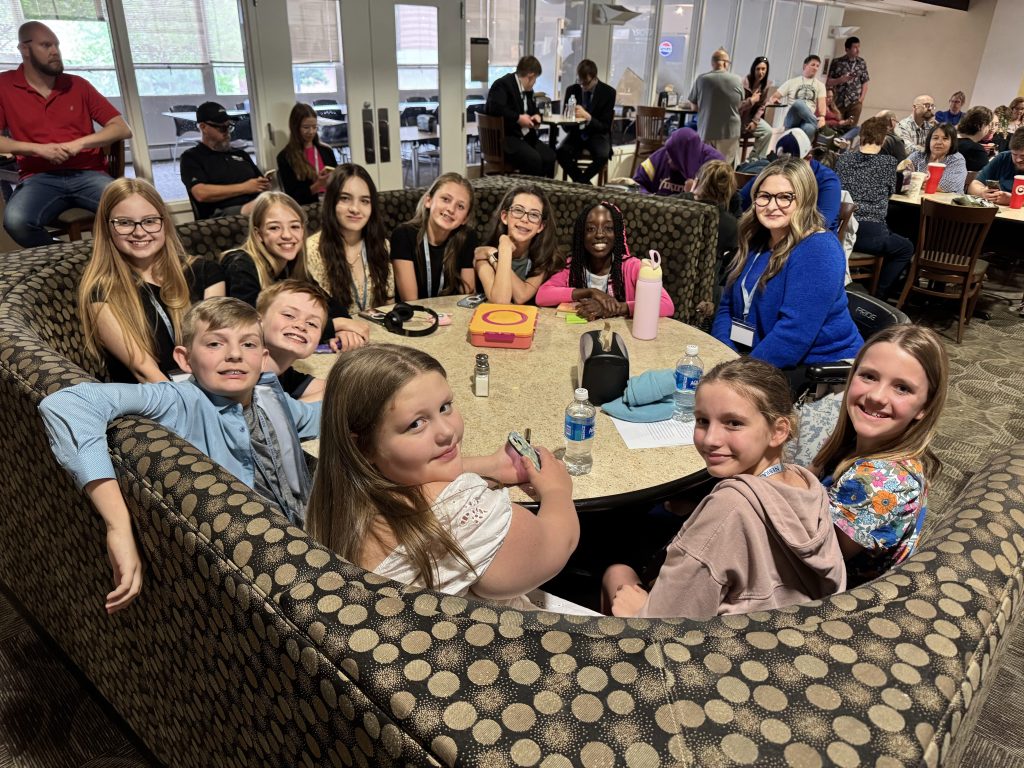 A group of children and teenagers sitting around a circular table in a casual dining setting, smiling and interacting with each other.