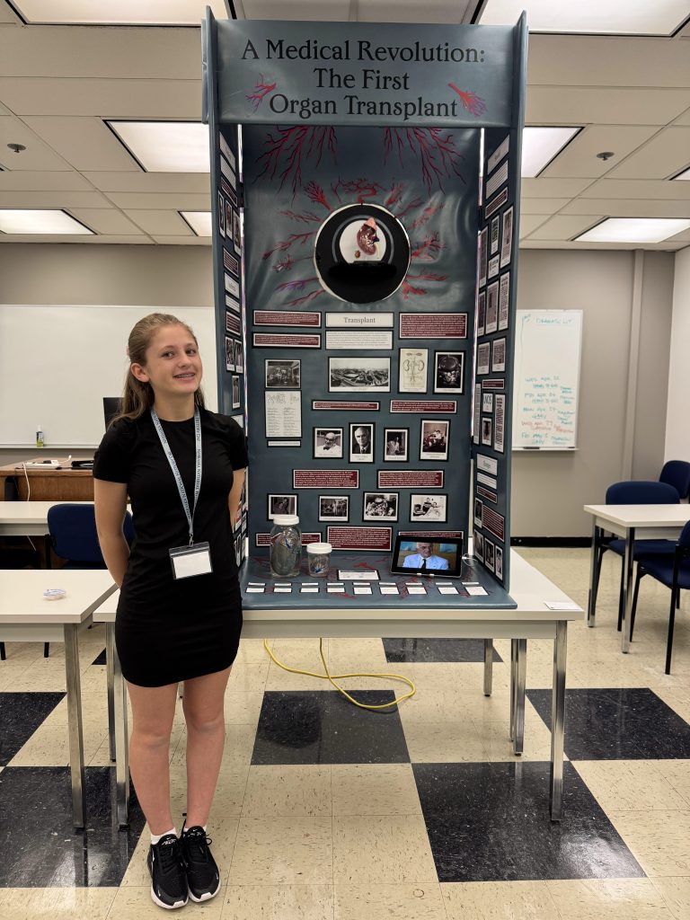 A student stands beside a display board titled 'A Medical Revolution: The First Organ Transplant,' featuring various images and information related to organ transplantation.