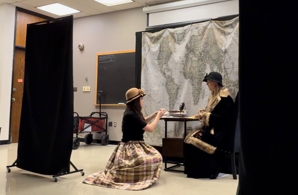 A scene depicting two women in period costume, one kneeling and one seated at a table in front of a map backdrop, in a classroom setting.