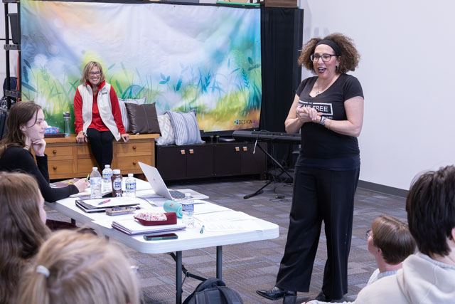 A speaker engages with an audience during a presentation, with a colorful backdrop. Two attendees listen attentively, while another participant sits at a table with notebooks and water bottles.