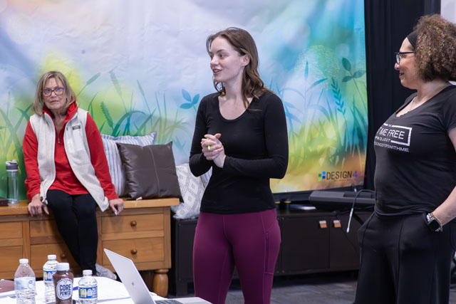 A woman speaking engagingly in front of a group, with two seated women listening. The background features a colorful nature-themed mural, and there are laptops and water bottles on a table.