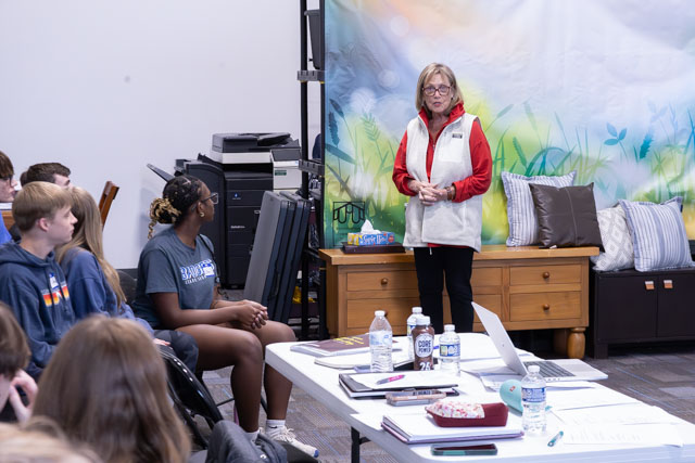 A woman in a red and white vest speaks to a group of students seated in a classroom setting, with a colorful backdrop and various items on a table.