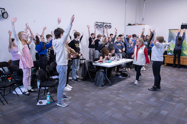A group of students in a classroom raising their hands, engaged in an interactive activity. Some are seated at tables while others stand with arms raised, creating a lively atmosphere.