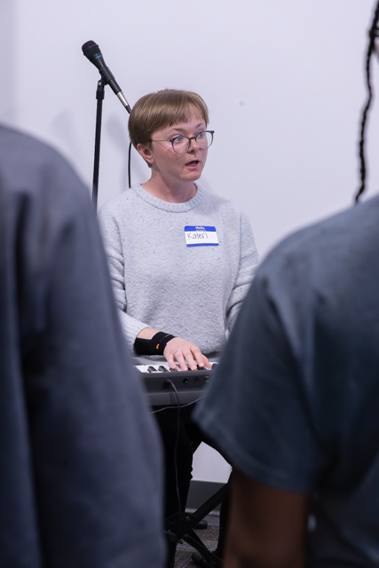 Person playing a keyboard while looking surprised or focused, wearing a gray sweater and glasses, with a name tag visible; microphone stand in the background.