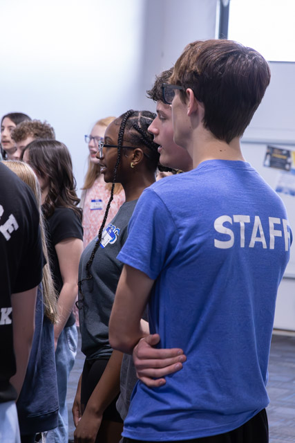 A group of young people standing in a line, with one individual wearing a blue STAFF shirt. They appear engaged and are facing forward, indoors with bright lighting.