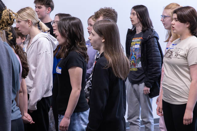 A group of diverse young people standing in a line, engaged in an activity, possibly a rehearsal or practice. They are facing forward, wearing casual clothing, and some are wearing name tags.