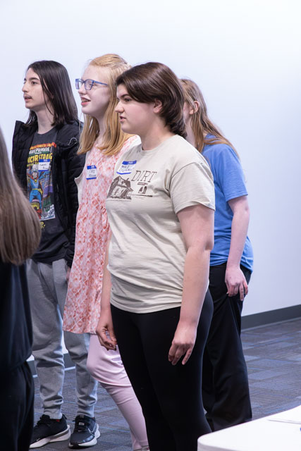 A group of teenagers standing in a line indoors, some wearing casual clothing and name tags, engaging in an activity.