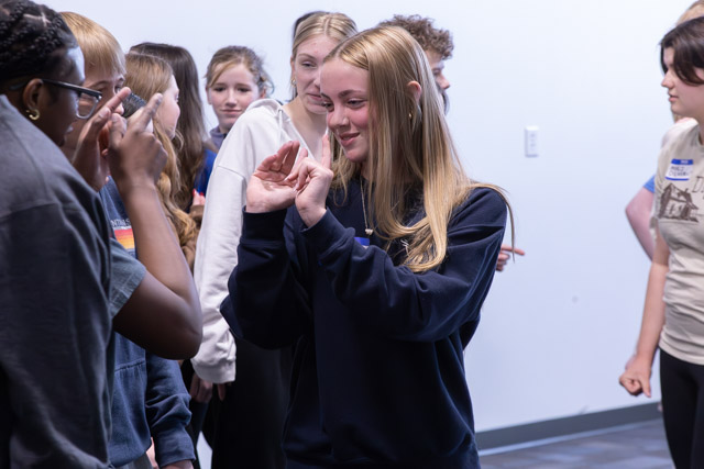 A group of teenagers participating in an interactive activity, with one girl in the foreground gesturing with her hands and smiling. Several peers are engaged in the background.