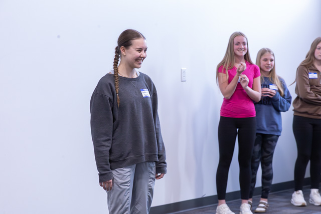 A group of four girls in a light-filled room, with one girl in the foreground wearing a gray sweatshirt and smiling. The other three girls, standing behind her, are clapping and smiling as well.