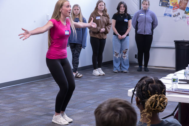 A woman in a pink top performs energetically in front of a group of seated young people during a workshop or presentation.